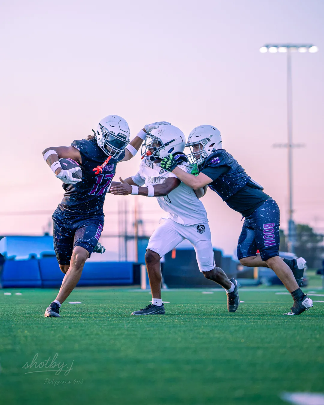 A WTX Boom running back in a dark number 17 jersey stiff-arms a defender's helmet, forcing him back as a teammate closes in, dusk sky behind them